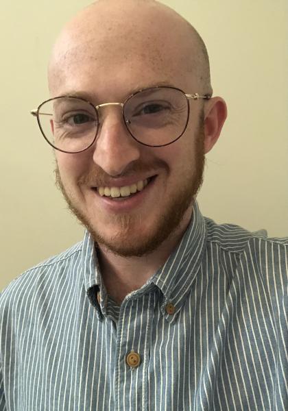 image of smiling white male with glasses and a reddish beard. Raymond is wearing a blue and white striped collared shirt 