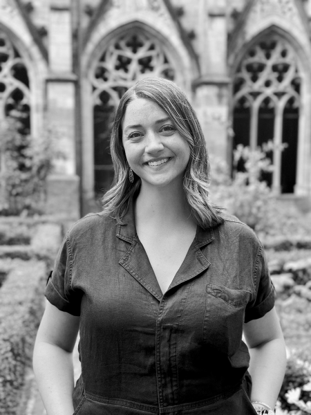 A black and white portrait of Leah smiling at the camera, with blurred gothic architecture in the background.