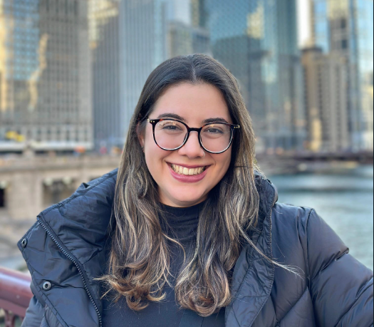 A photo of Laura Vieira, a white-looking woman with long brown hair and blond highlights. She has big round glasses and a smile. She wears dark red lipstick and a black puffer jacket. Behind her are tall buildings in Chicago.