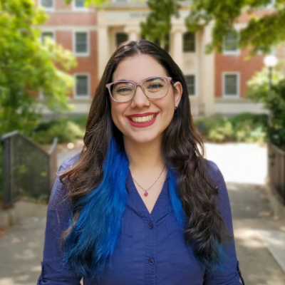 A photo of Laura Vieira, a white woman wearing glasses, a dark blue shirt and dark pink lipstick. She has a small silver necklace with a pink stone, and blue streaks on her hair. Behind her there are a few trees and the front of Gilbert Hall.
