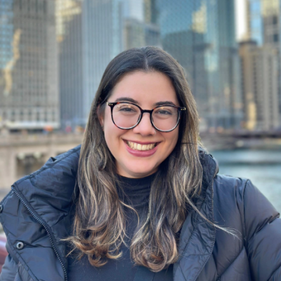 A photo of Laura Vieira, a white-looking woman with long brown hair and blond highlights. She has big round glasses and a smile. She wears dark red lipstick and a black puffer jacket. Behind her are tall buildings in Chicago.